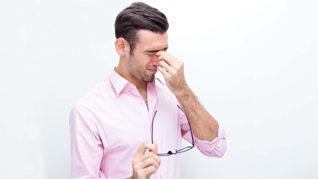 A man with eye discomfort touches his eyes with one hand and holds his glasses with the other.