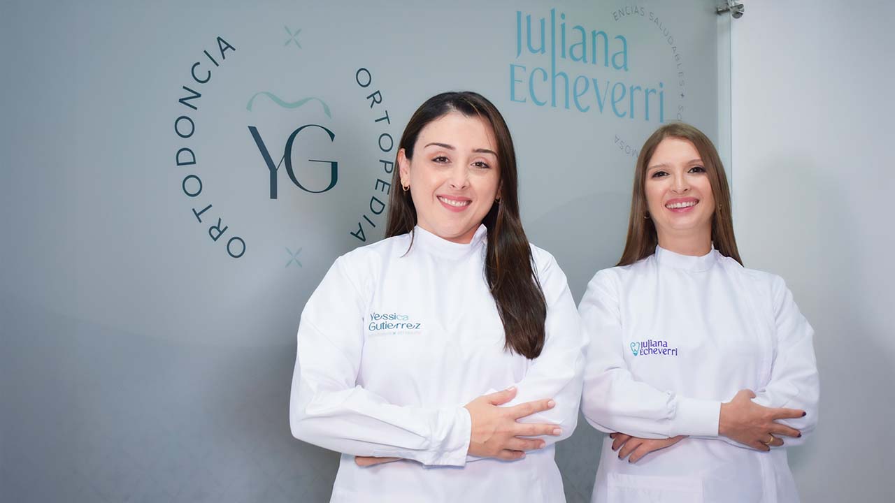 Dos odontólogas posan frente a la pared donde se ve el logo de su marca personal.