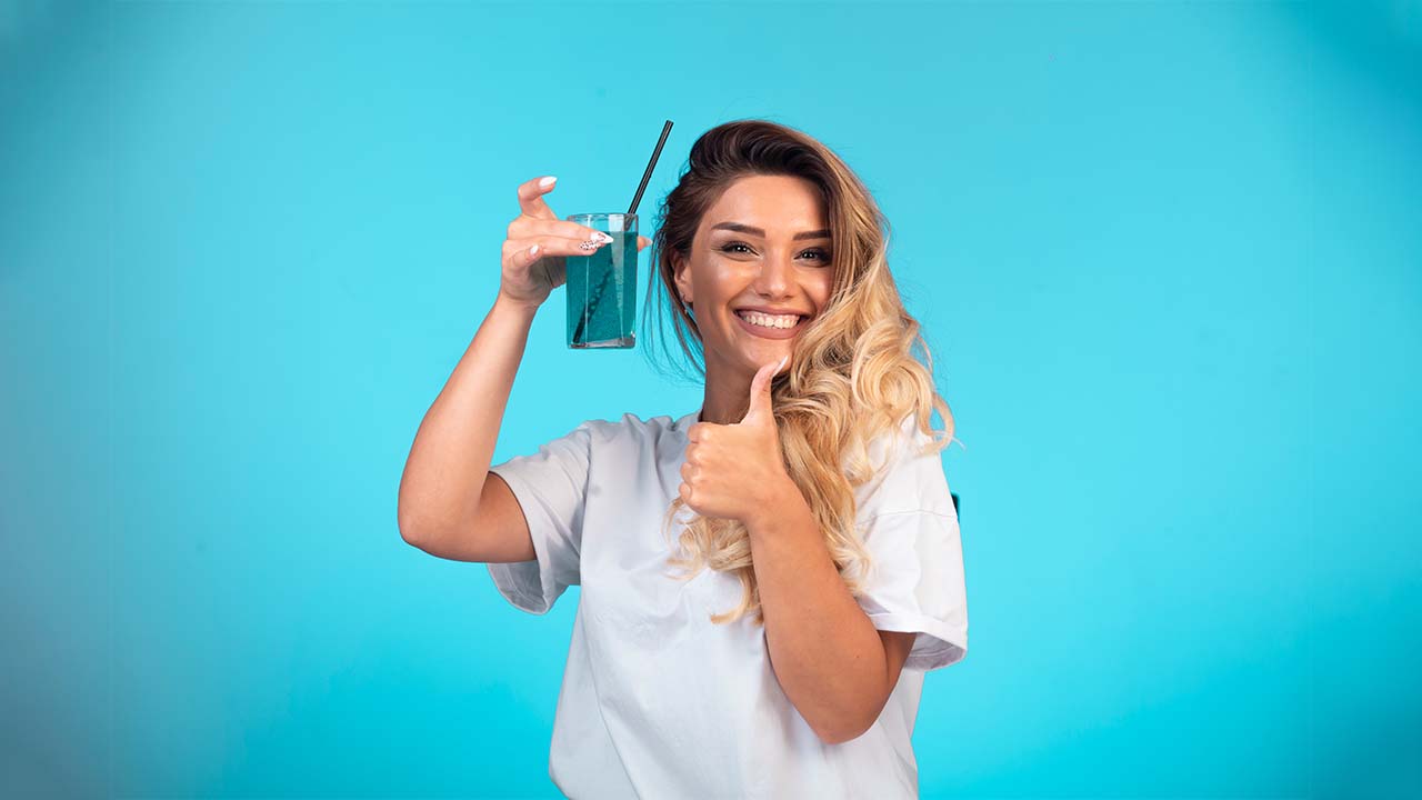 Una mujer sostiene un remedio casero en una mano y nos da una señal de éxito con la otra mientras mira a la cámara con una risa.