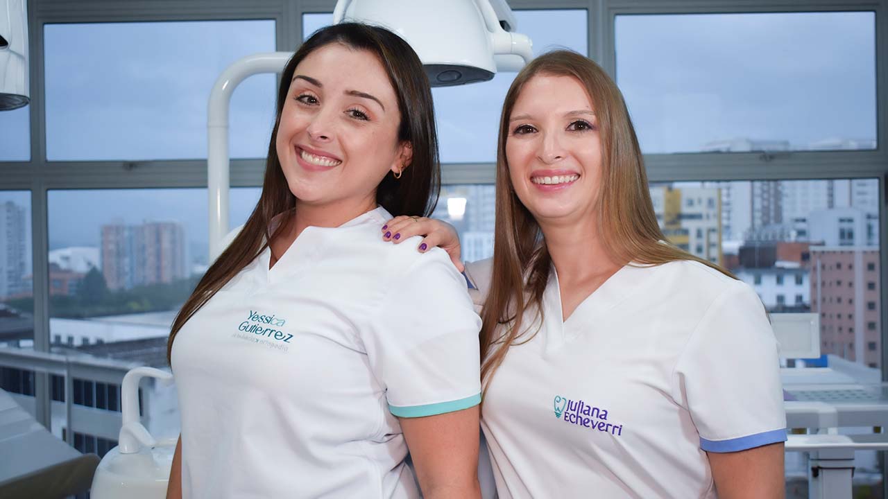 Dos odontólogas sonriendo ante la cámara desde su consultorio.